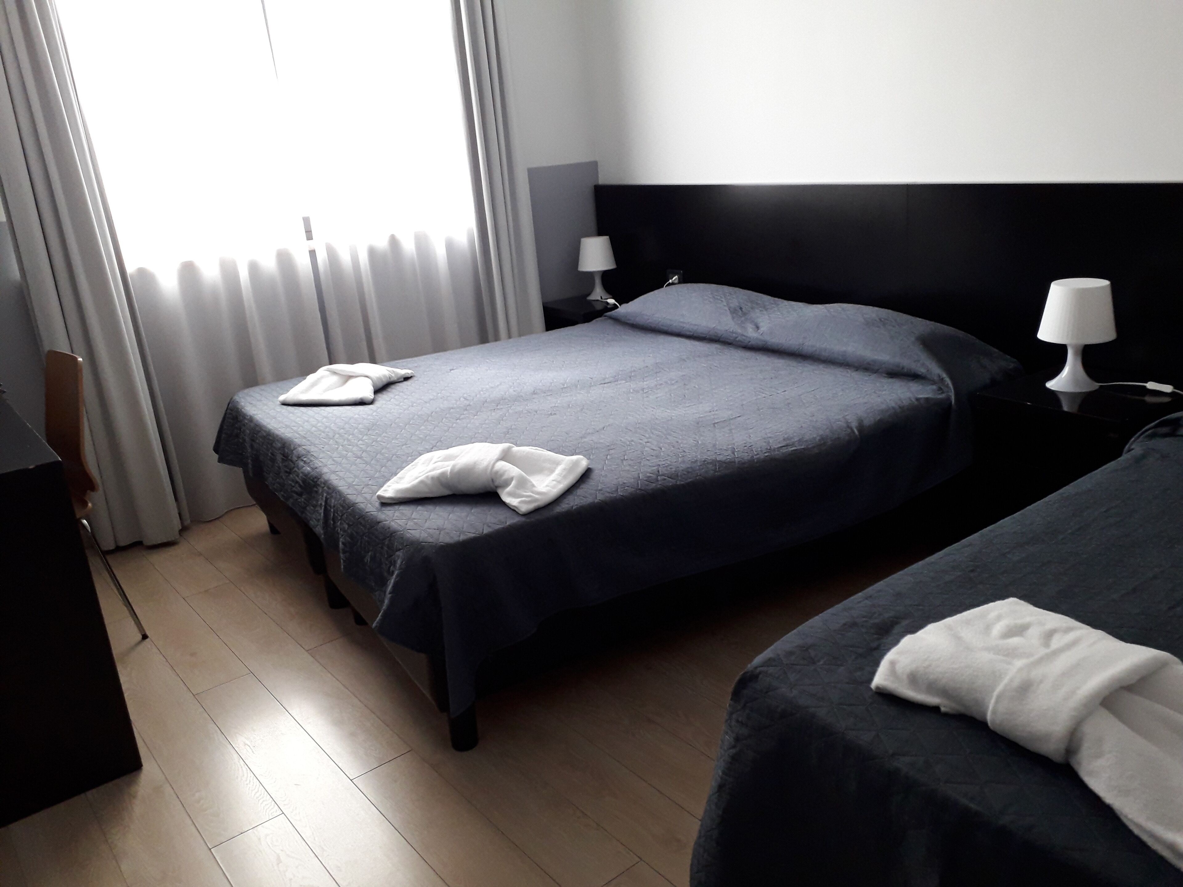 Quarto de hotel com duas camas, uma mesa de cabeceira com abajur e vista para a cidade pela janela.