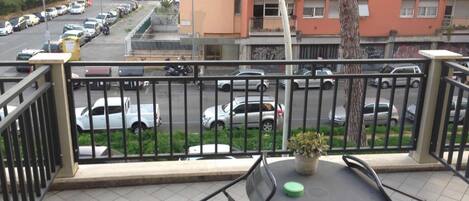 Un balcone con tavolo e sedie, affaccia su una strada con auto parcheggiate e un edificio con balconi.