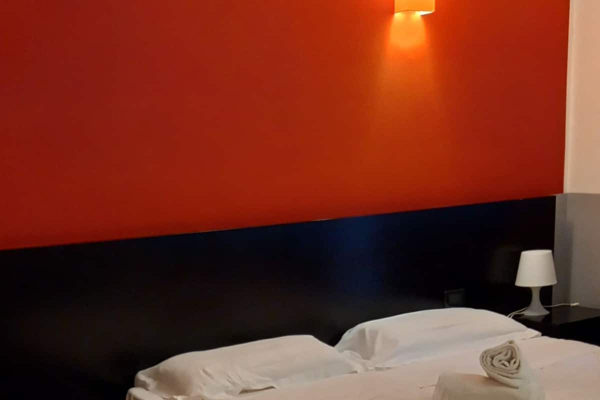 A hotel room with two beds, a red accent wall, a white lamp, and a wooden floor.