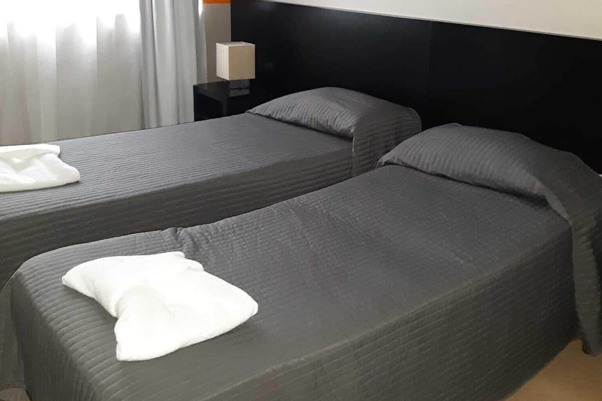 A hotel room with two beds, a nightstand, a lamp, and a window with curtains.