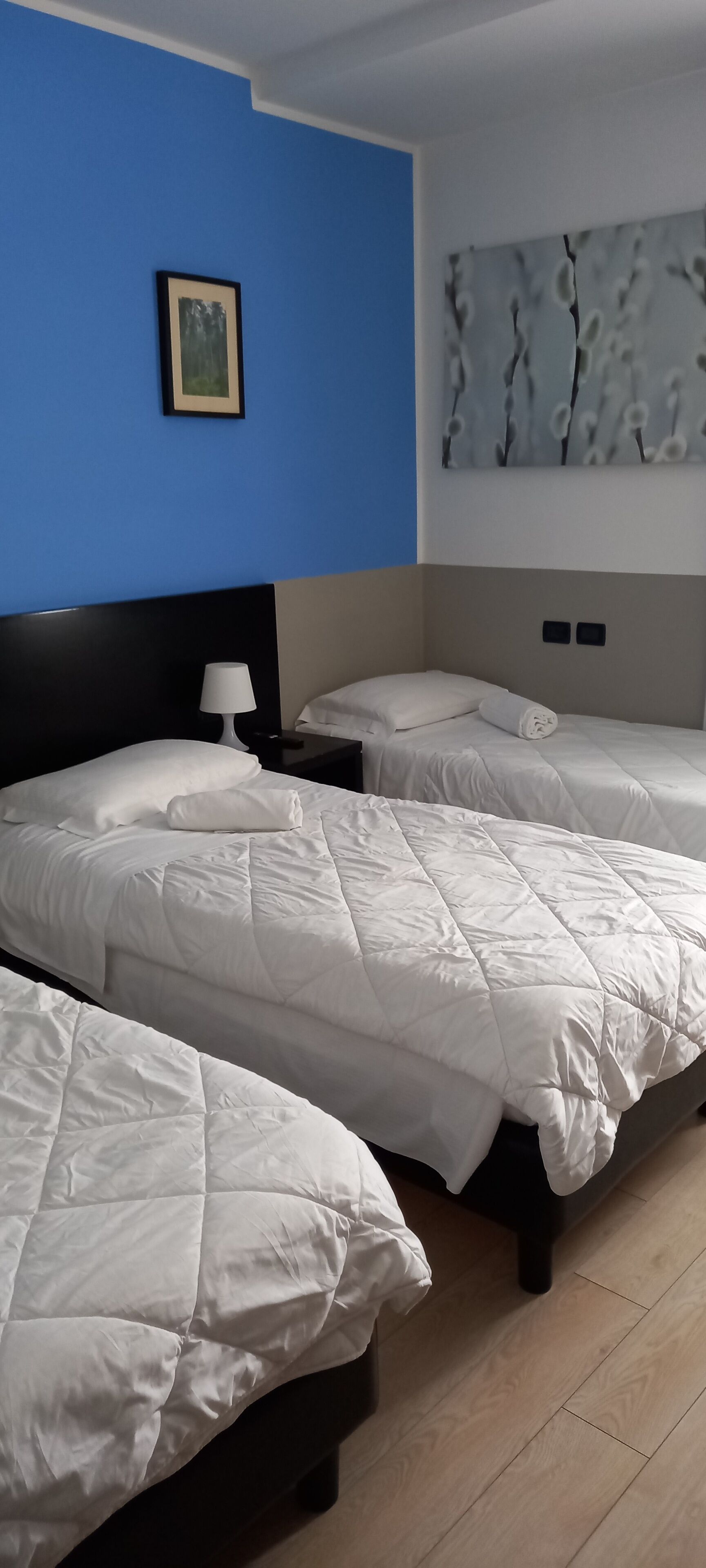 A hotel room with two beds, a nightstand with a lamp, a framed picture on the blue wall, and a large abstract painting above the beds.