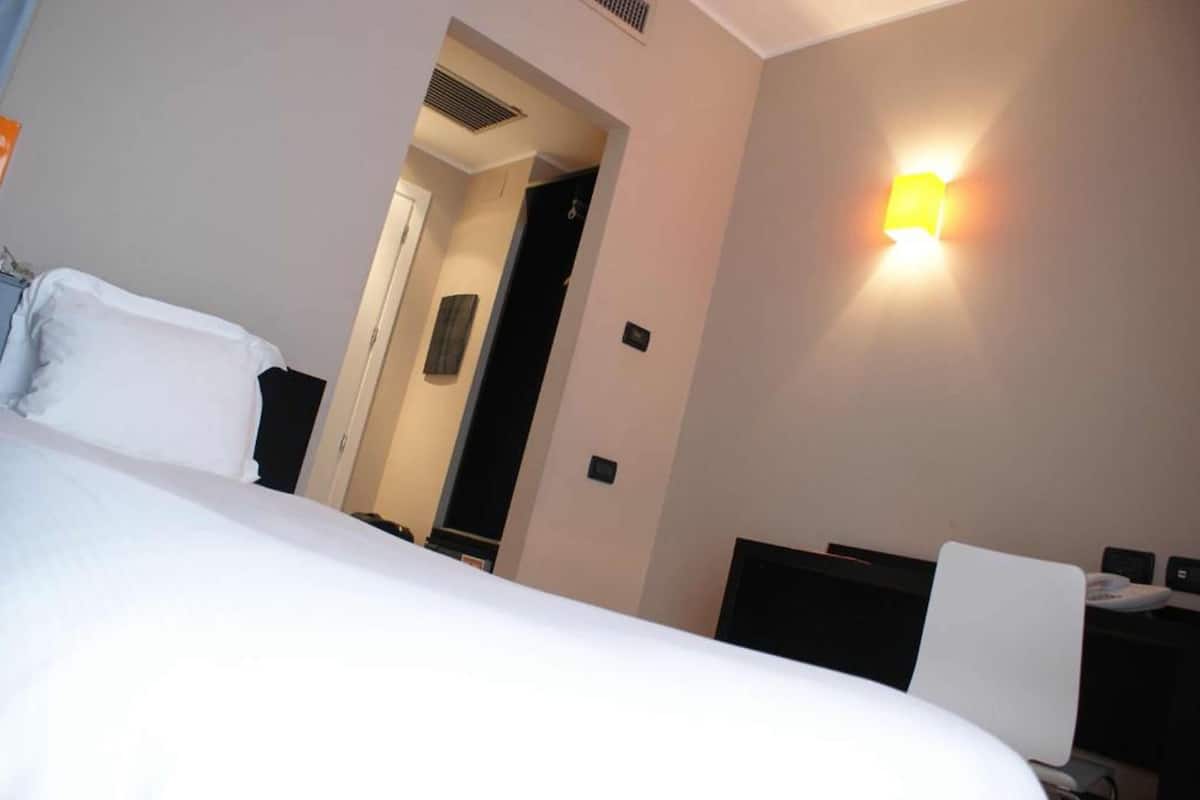 A hotel room with a bed, a desk, and a wall-mounted light fixture.