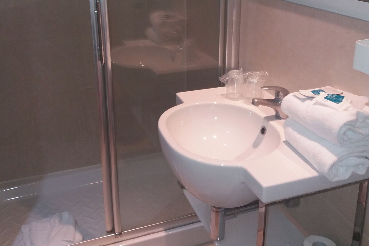 A bathroom with a glass shower enclosure, a white sink with a mirror above it, and a toilet with a roll of toilet paper on the floor.