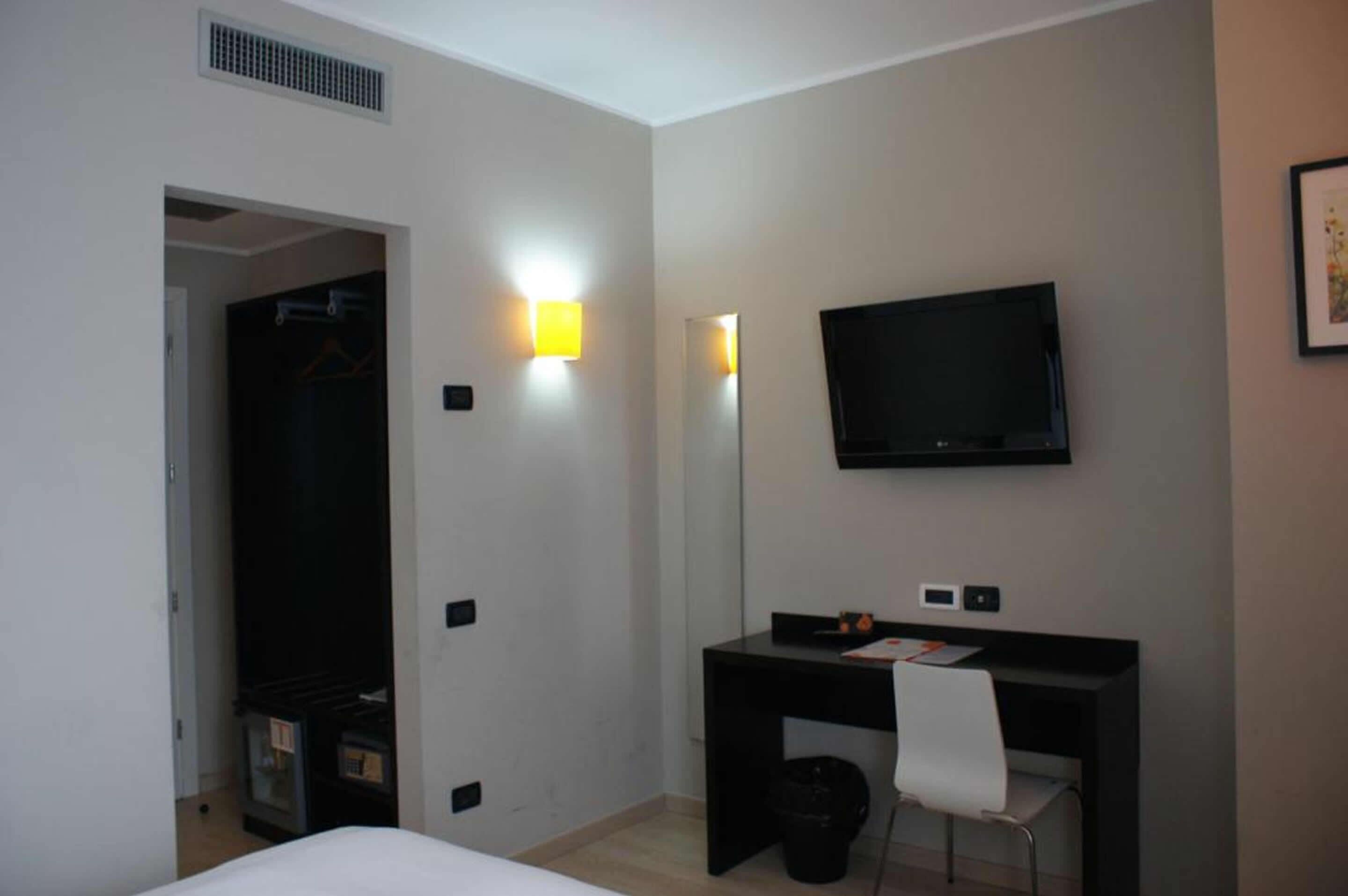 A hotel room with a bed, a desk with a chair, a television mounted on the wall, and a door leading to a closet.