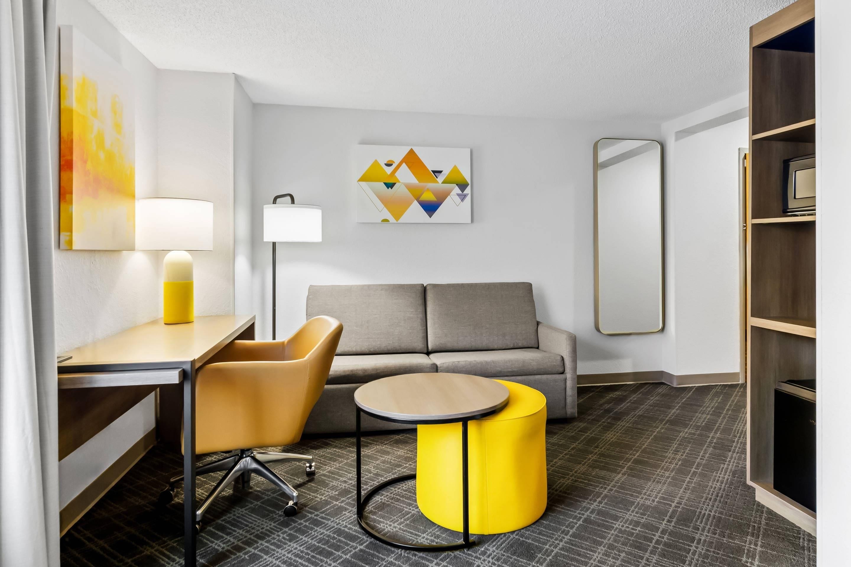 Una habitación de hotel moderna con un sofá, una mesita redonda pequeña, una silla, una lámpara, un cuadro y un espejo.