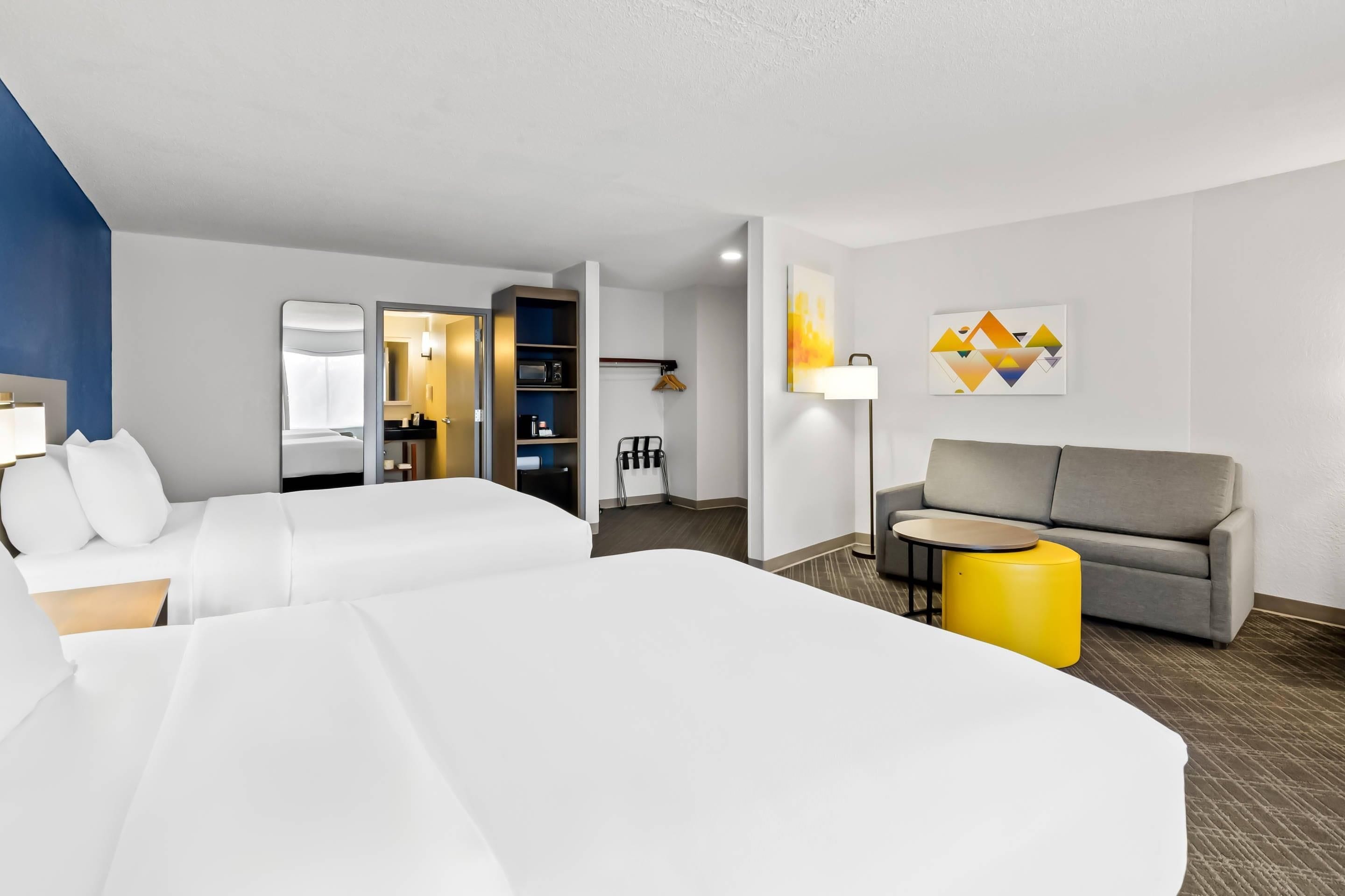 Une chambre d’hôtel avec deux lits, un canapé, une petite table et un pouf jaune.