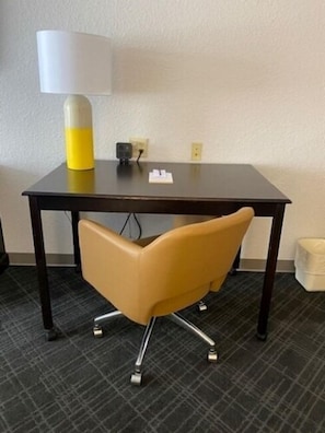 Een hotelkamer met een donker houten bureau, een geel-witte lamp en een moderne stoel.
