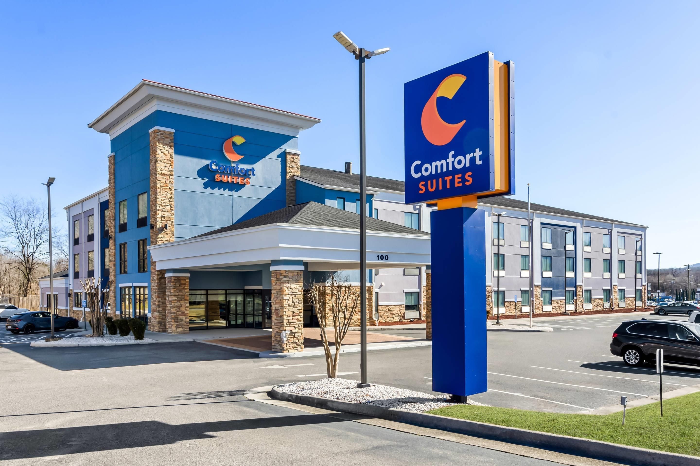 Un hotel Comfort Suites con un diseño moderno, que presenta un logotipo azul y naranja llamativo, un amplio estacionamiento y un exterior bien cuidado.