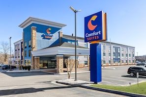 Un hotel Comfort Suites con un diseño moderno, que presenta un logotipo azul y naranja llamativo, un amplio estacionamiento y un exterior bien cuidado.