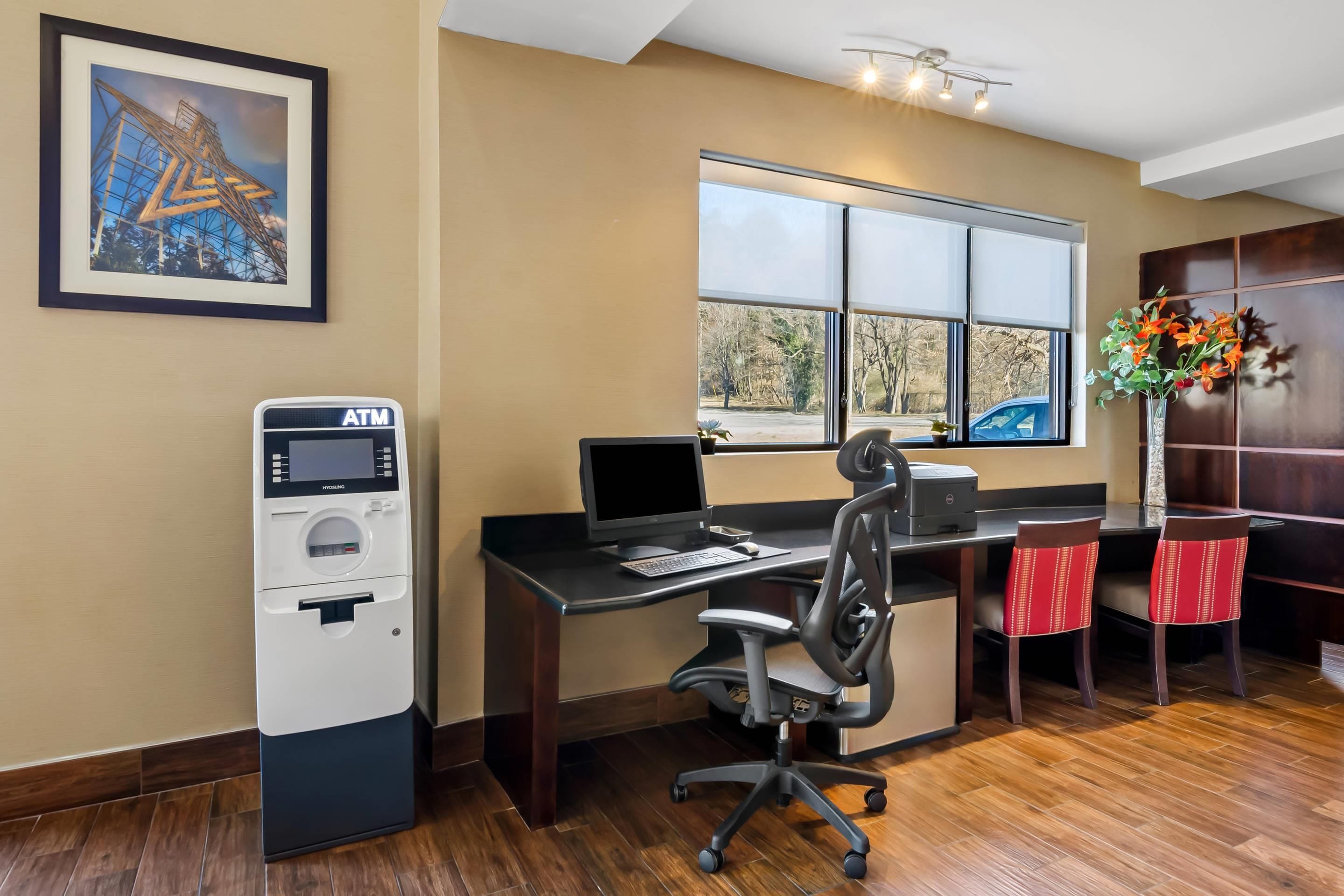 A hotel room with a desk, computer, chair, ATM, window with view, and a framed picture on the wall.