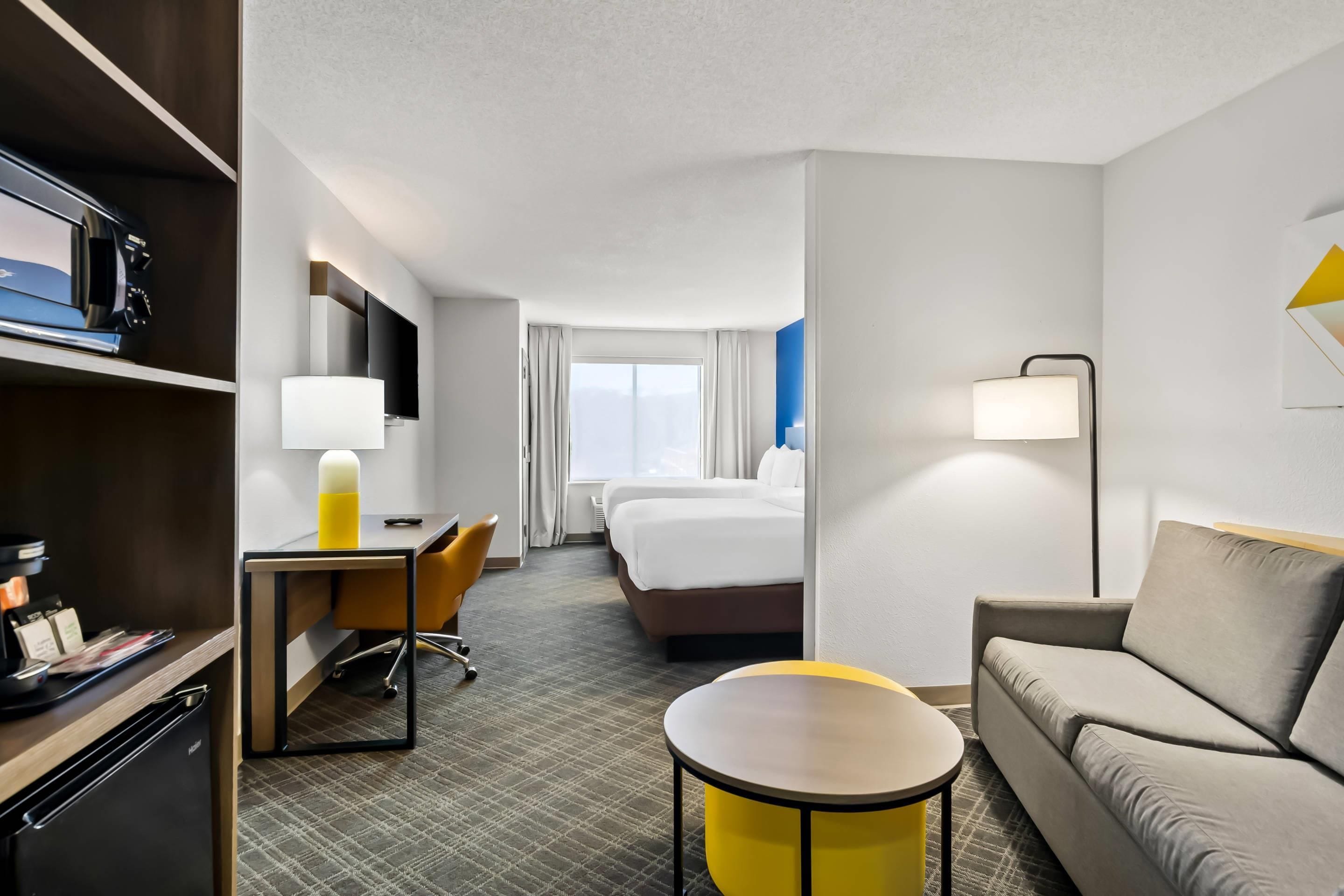 Une chambre d’hôtel comprenant un lit, un bureau, une chaise, un canapé, une petite table, une télévision et un mini-bar.