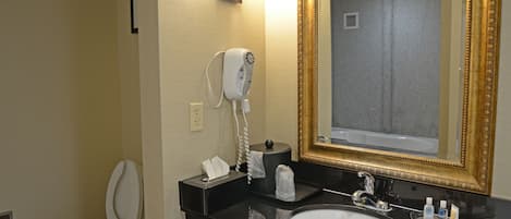 Un baño con inodoro blanco, lavamanos con encimera negra, espejo con marco dorado y un secador de pelo montado en la pared.
