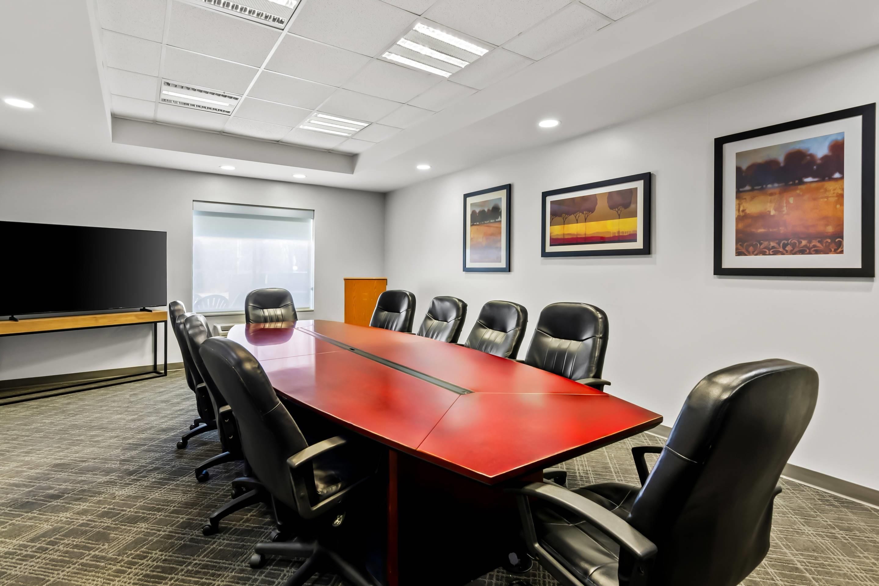 A conference room with a long red table, black chairs, a large flat-screen TV, and framed artwork on the walls.