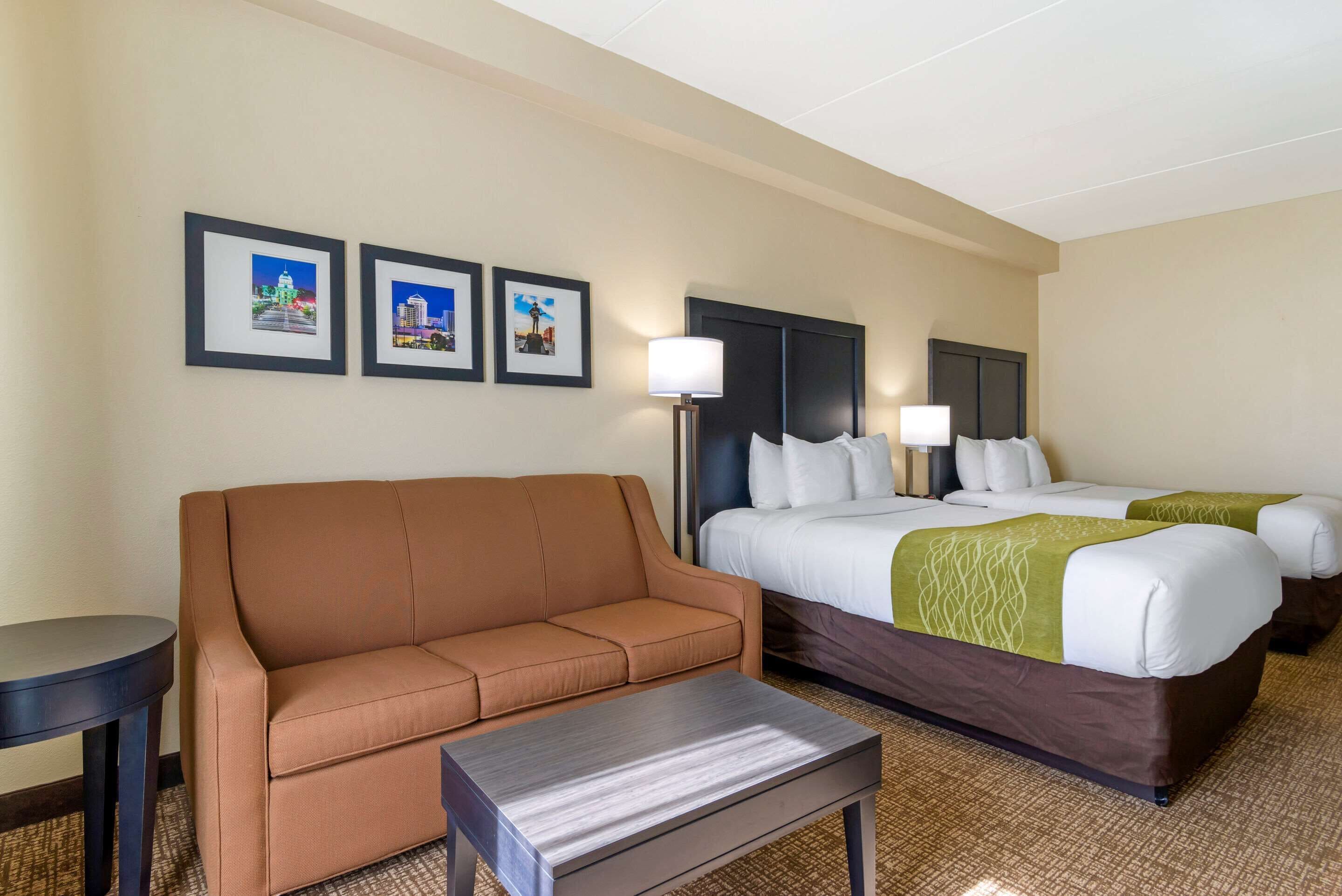 A hotel room with a brown sofa, a small table, two beds with white and green bedding, and three framed pictures on the wall.
