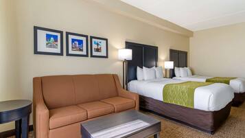 A hotel room with a brown sofa, a small table, two beds with white and green bedding, and three framed pictures on the wall.