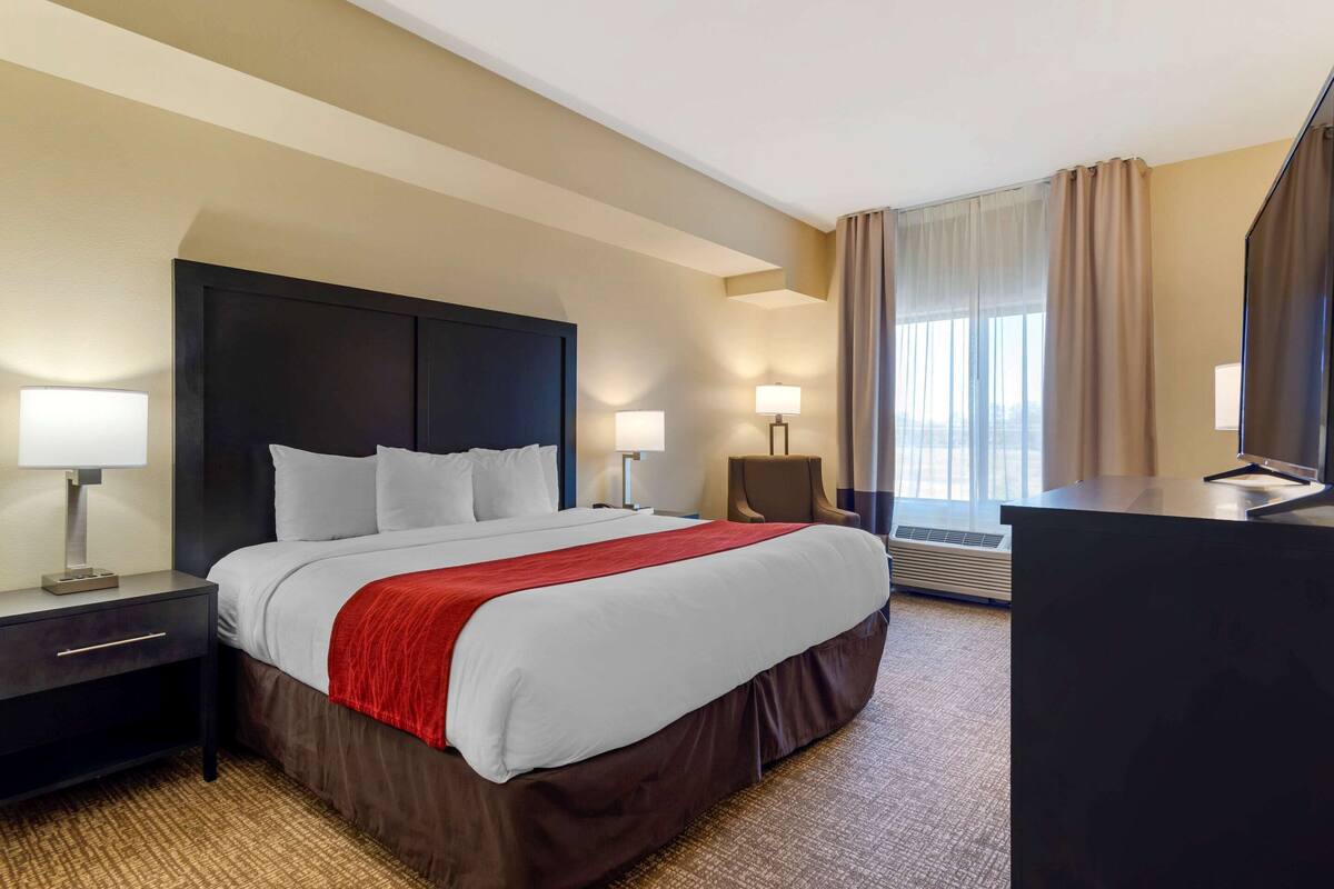 A hotel room with a large bed, a desk, two bedside lamps, a chair, and a large window with curtains.