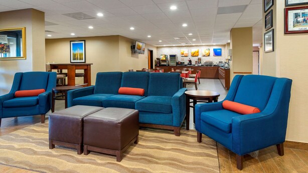 A hotel lobby with blue armchairs, a sofa, and a coffee table. There is a reception desk and a kitchen area in the background. - Comfort Inn & Suites Montgomery Eastchase (Montgomery)