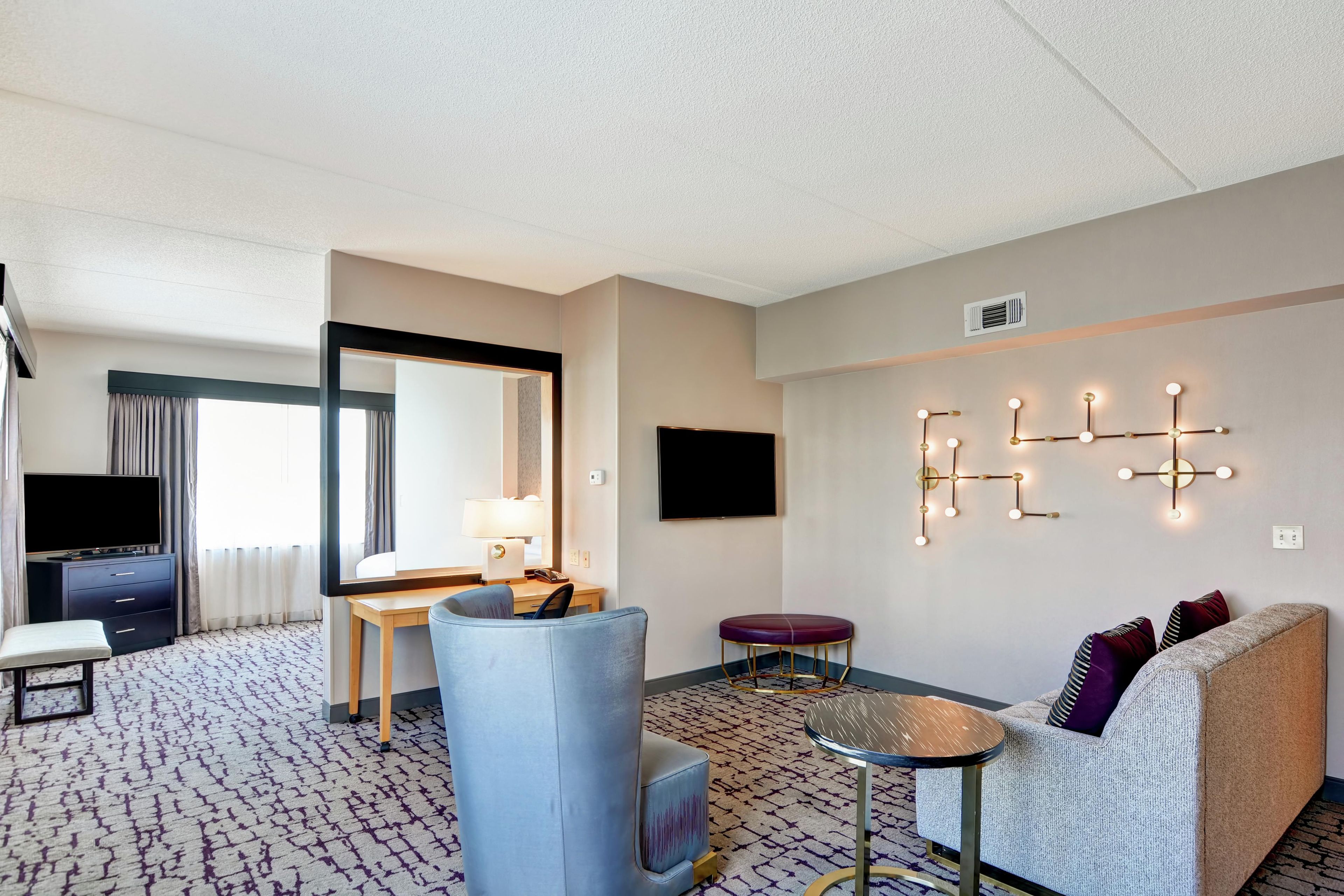 Habitación de hotel con cama, escritorio, silla, sofá, mesita pequeña, espejo y lámpara fijada en la pared.