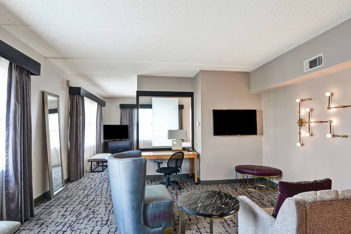 A hotel room with a desk, chair, TV, and seating area.