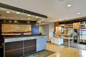 A hotel reception area with a modern design, including a reception desk, a large artwork, and a kitchenette area with a refrigerator and a coffee machine.