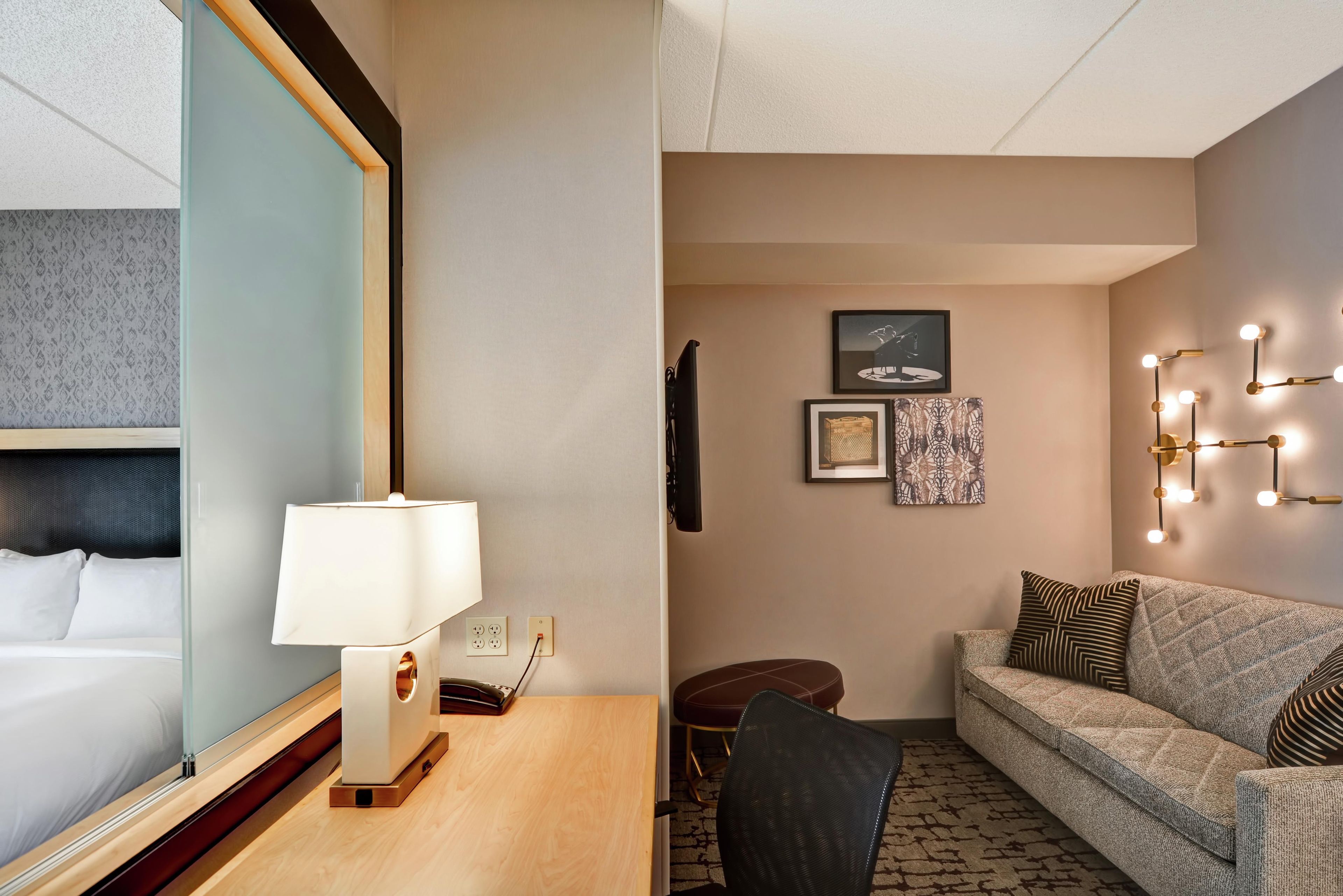 A hotel room with a bed, a desk with a lamp, a sofa, and a wall-mounted light fixture.