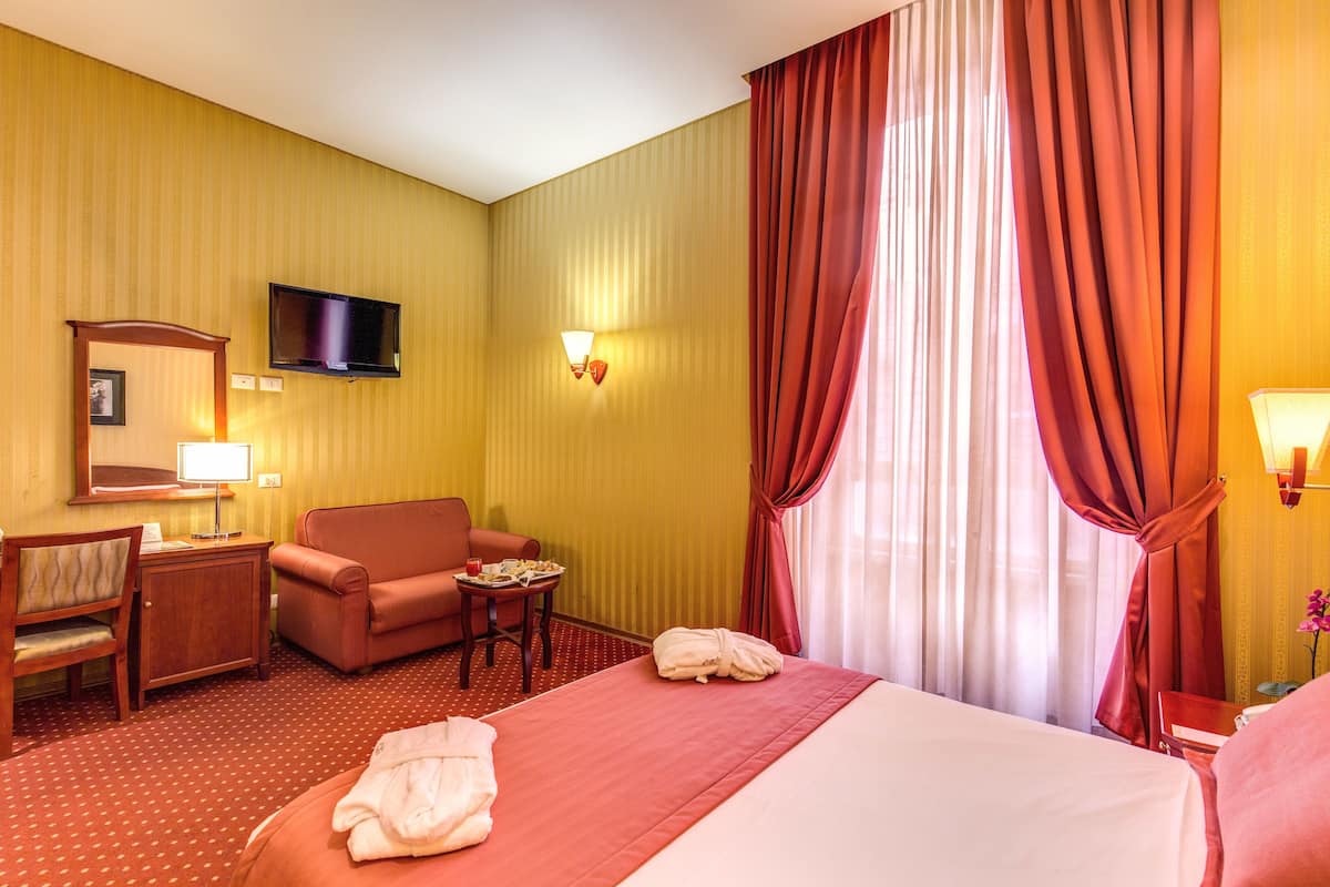 Ein Hotelzimmer mit Bett, Schreibtisch, Stuhl, Fernseher und einem Fenster mit Vorhängen.