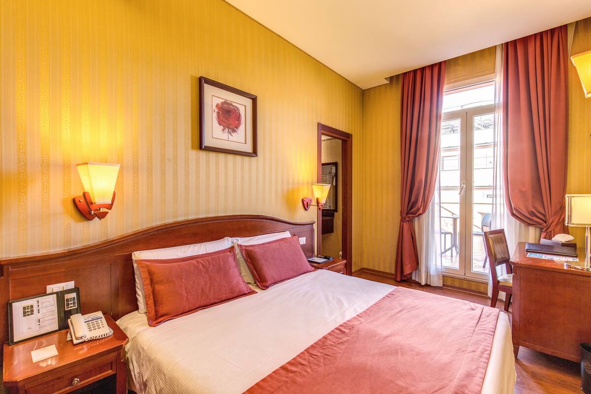 Ein Hotelzimmer mit einem großen Bett, einem Schreibtisch, einem Stuhl, einem Telefon und einem Fenster mit Vorhängen.
