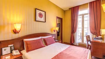 A hotel room with a large bed, a desk, a chair, a telephone, and a window with curtains.