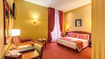 A hotel room with a bed, a desk with a lamp, a chair, a small table with a tray, and a picture on the wall.