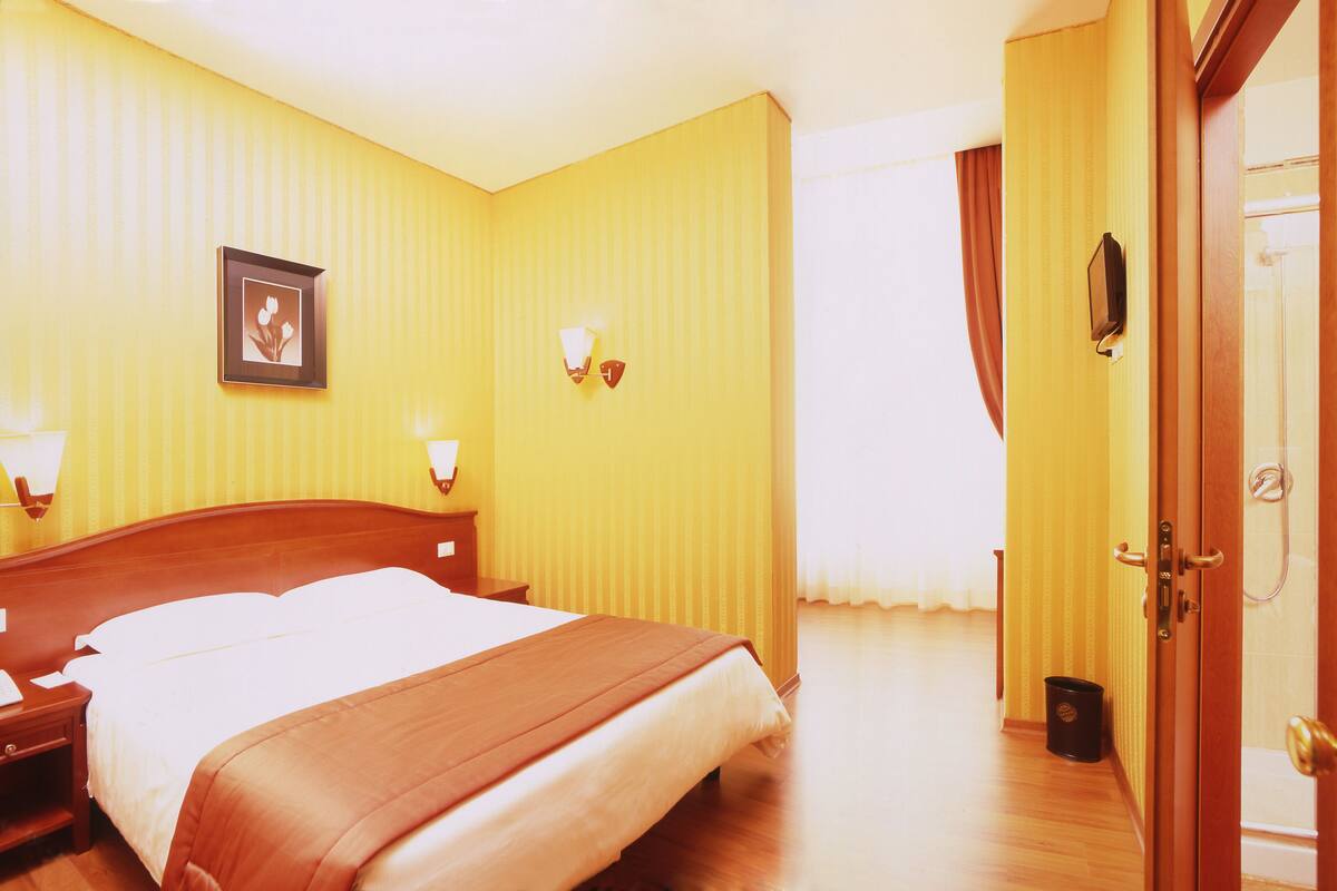 Ein Hotelzimmer mit Bett, Nachttischlampen, einem gerahmten Bild an der Wand und einer Tür rechts.