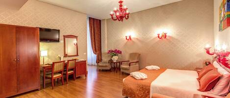 A hotel room with a wooden floor, a bed with orange bedding, a desk with a chair, a television, and a chandelier.