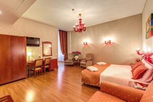 A hotel room with a wooden floor, a bed with orange bedding, a desk with a chair, a television, and a chandelier.