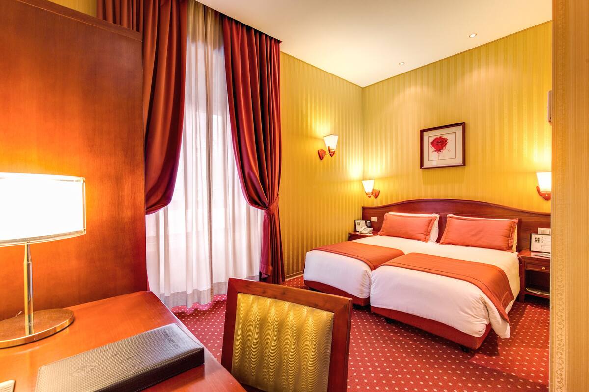 Ein Hotelzimmer mit zwei Betten, einem Schreibtisch mit Lampe, einem Stuhl und einem großen Fenster mit Vorhängen.