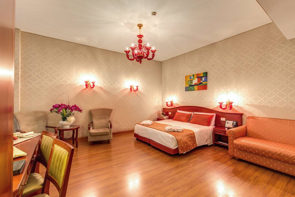 Ein Hotelzimmer mit Holzboden, einem Bett mit roter Bettwäsche, einer Couch, einem Schreibtisch mit Stuhl und einer Kandelaber.
