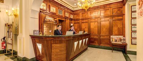 A reception desk with two staff members, a chandelier, and a clock on the wall.