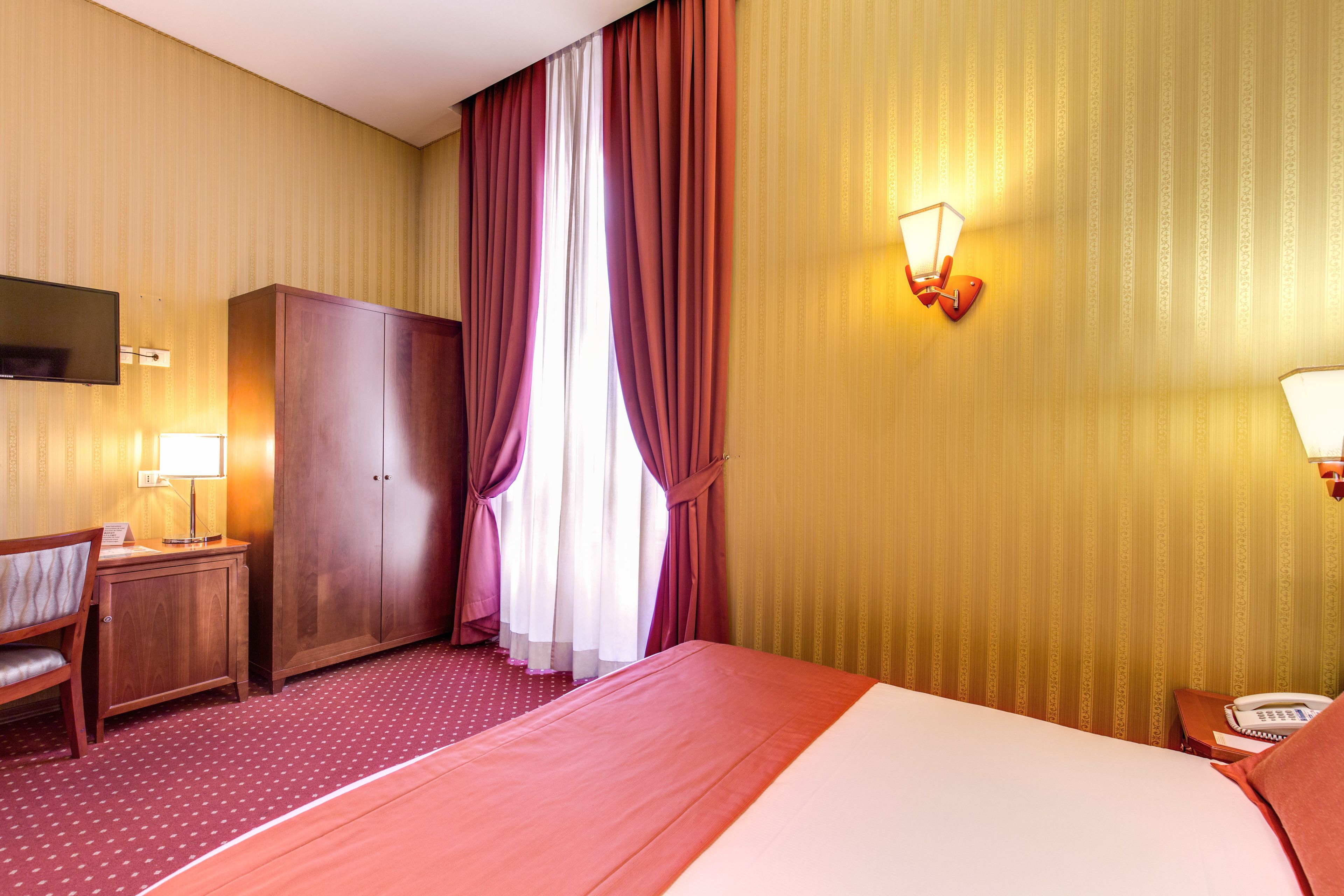 A hotel room with a bed, a desk, a wardrobe, and a wall-mounted lamp.