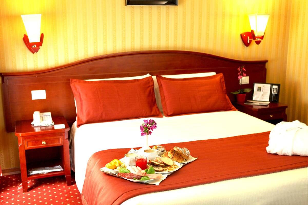 Ein Hotelzimmer mit einem ordentlich bezogenen Bett, einem Nachttisch mit Lampe und einem Tablett mit Frühstücksangeboten auf dem Bett.