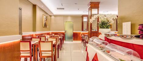 A hotel dining area with wooden tables and chairs, a buffet line with food covers, and a reception desk.