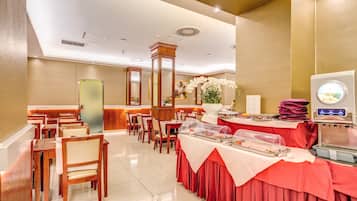 Espaço de refeições de hotel com várias mesas e cadeiras, um serviço de buffet e um pilar decorativo.