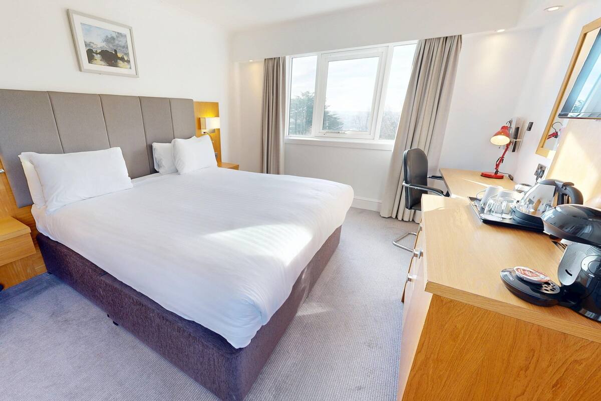 A hotel room with a large bed, a wooden desk, a chair, a desk lamp, a telephone, and a window with curtains.