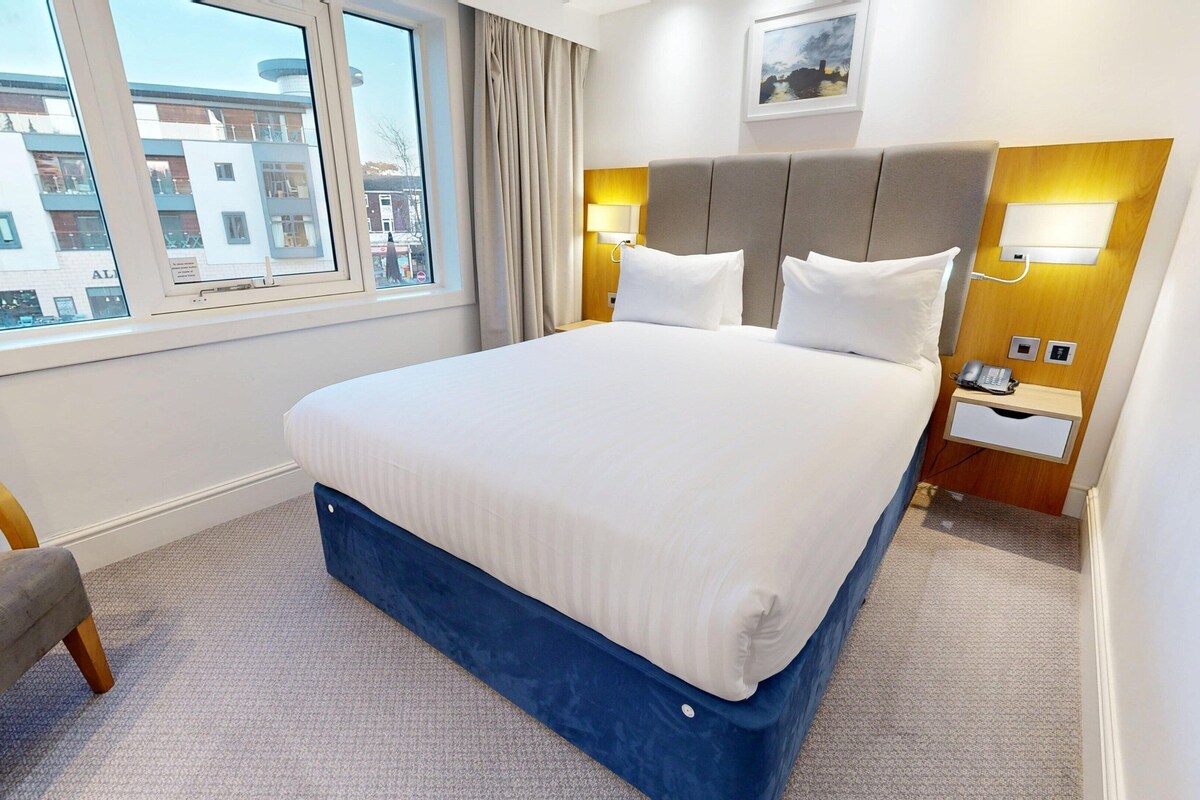 Habitación de hotel con una cama grande, una mesita de noche con teléfono, una silla y una ventana con vistas a edificios.