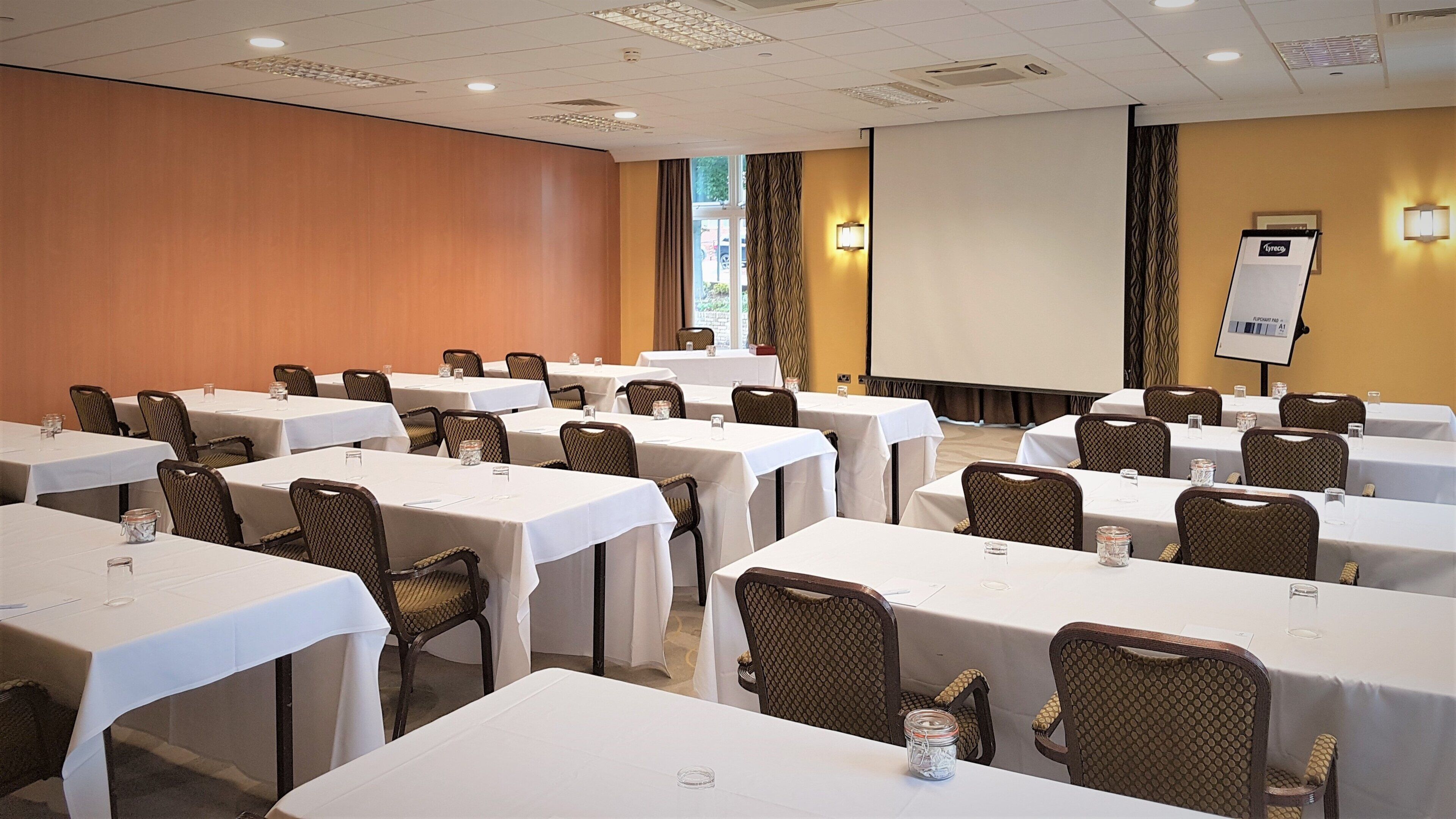 Un salón de conferencias con varias mesas, sillas, una pantalla de proyector y una pizarra para presentaciones.