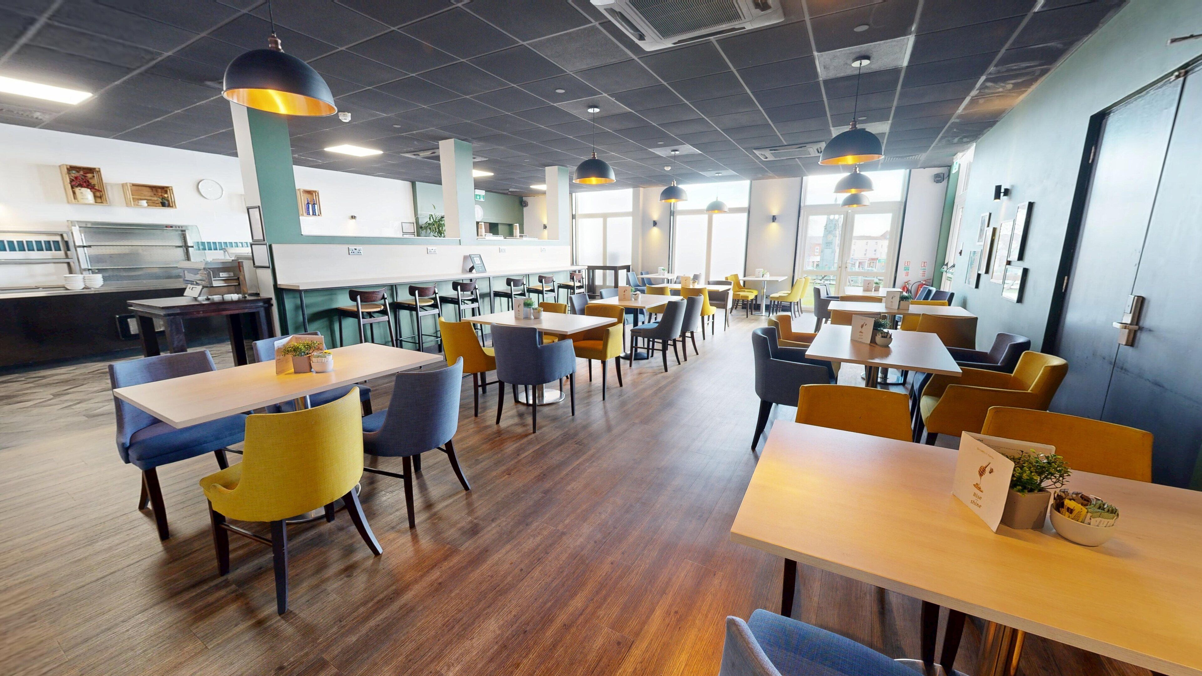 A spacious dining area with multiple tables and chairs, a kitchen in the background, and large windows allowing natural light.