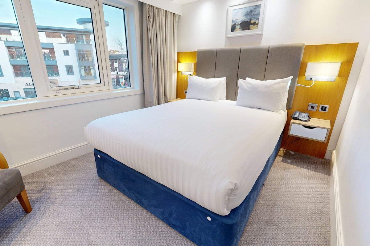 A hotel room with a large bed, a bedside table with a phone, a chair, and a window with a view of buildings.