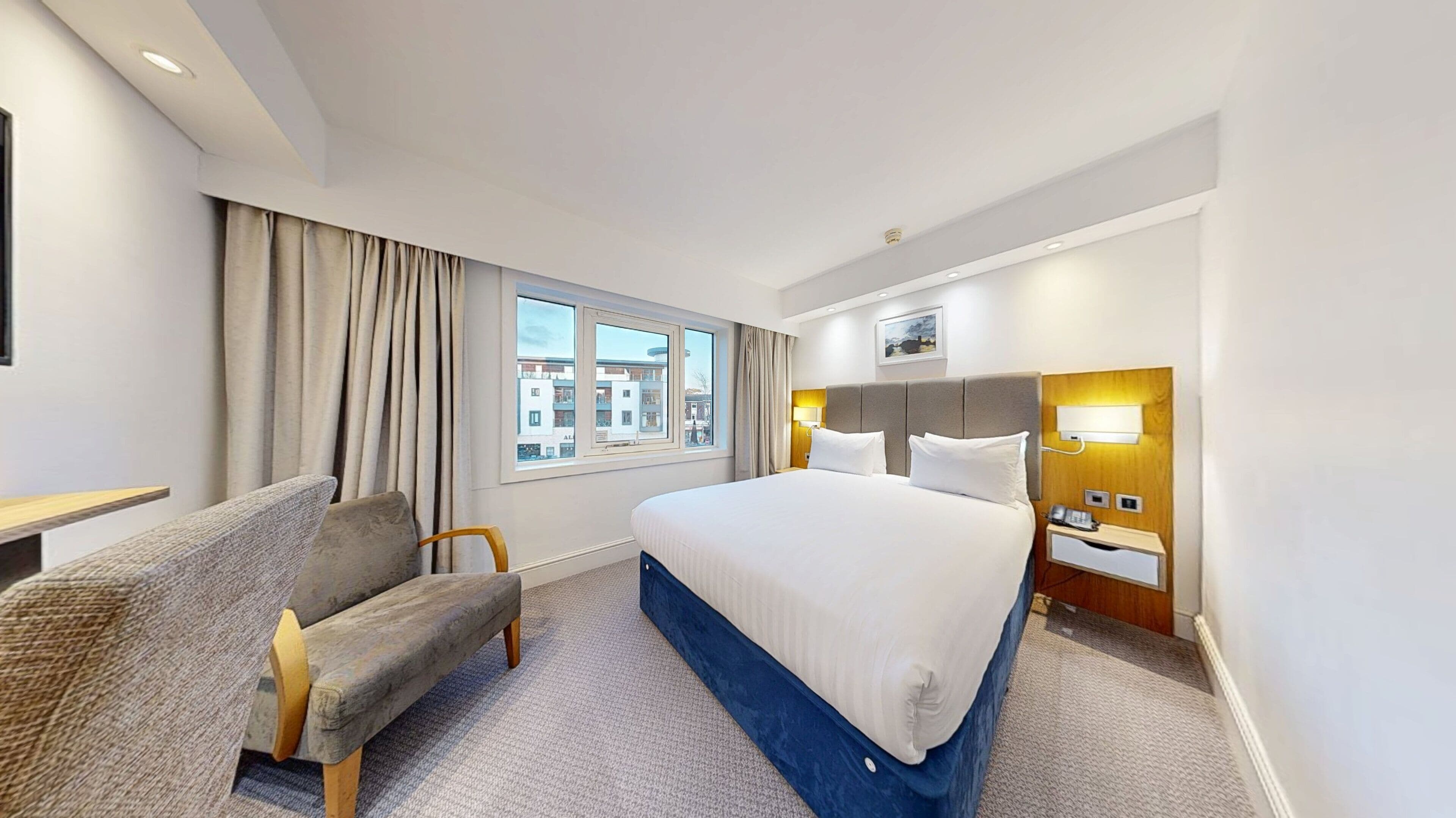 A hotel room with a bed, a desk, a chair, a window with curtains, and a view of buildings outside.