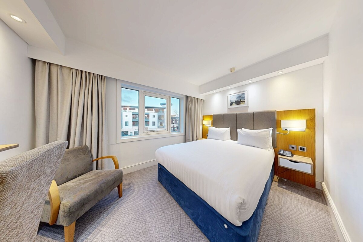 Habitación de hotel con cama, escritorio, silla, ventana con cortinas y vistas a edificios.