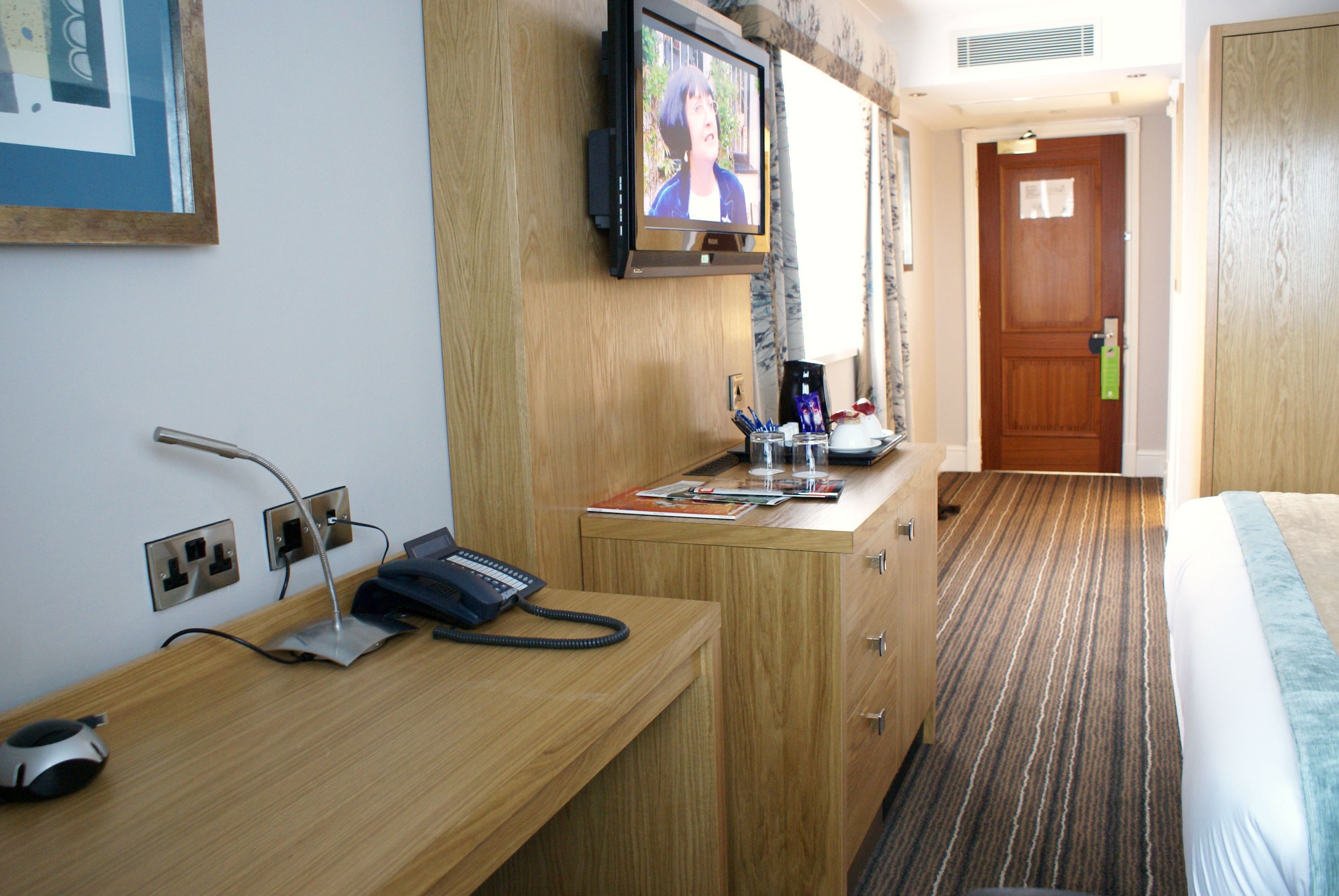 A hotel room with a wooden desk, a television mounted above it, a bed with striped bedding, and a door with a green handle.