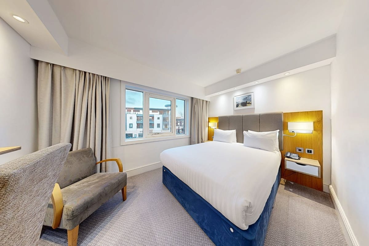 A hotel room with a bed, a desk, a chair, a window with curtains, and a view of buildings outside.