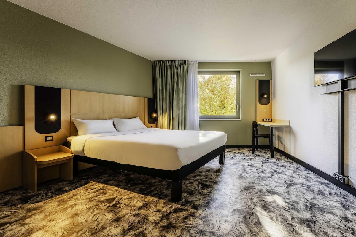Ein Hotelzimmer mit einem Bett, einem Nachttisch, einem Schreibtisch, einem Sessel, einem Fenster mit Vorhängen und einem am Wand montierten Fernseher.