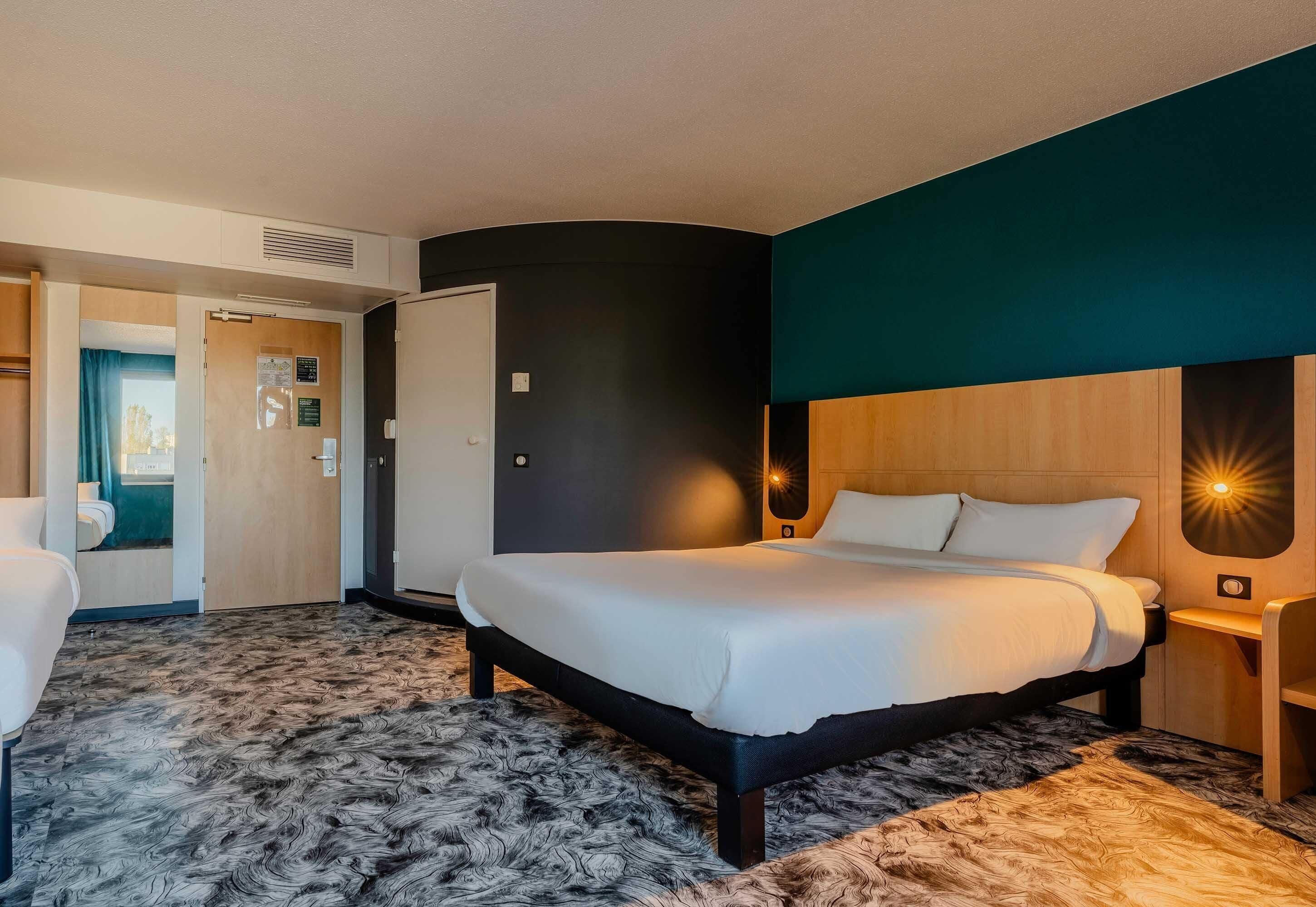 A hotel room with a bed, a desk, a chair, a door, and a wall-mounted light fixture.
